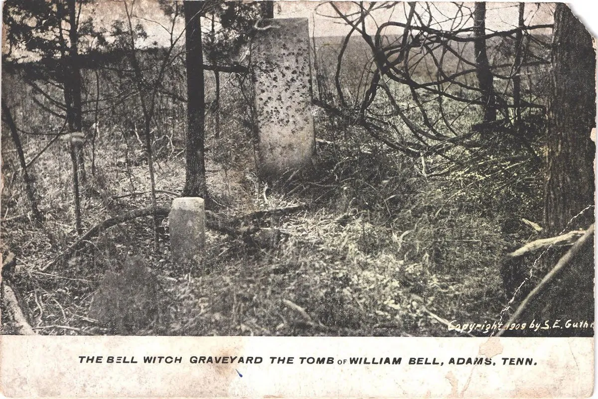 A colour-tinted photograph of a tall weathered gravestone and a short stone post among bare trees and undergrowth