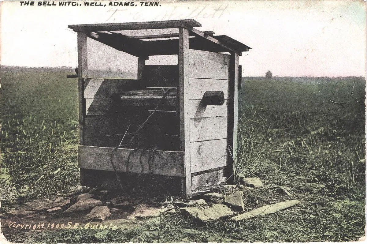 A colour-tinted photograph of a small wooden well house with a plank roof and a rope, standing alone in a flat grassy field