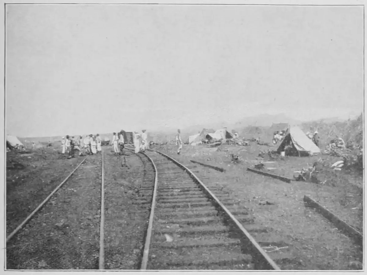 A black-and-white photograph of a railway line running through open, scrubby ground, with bell tents and a group of workmen beside the track in the distance